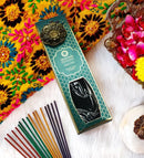 Eine türkis-goldene Schachtel Ayurveda Aqua Oud Classic Incense Sticks liegt diagonal auf einer weißen Oberfläche, umgeben von mehrfarbigen Räucherstäbchen. Die Szene ist mit einem leuchtenden, bestickten gelben Tuch und einer Silberschale mit roten und gelben Blütenblättern dekoriert.