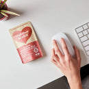 Eine Packung "together health ORGANIC VITAMIN B12" aus Shiitake-Pilz liegt auf einem weißen Schreibtisch neben einer weißen Computermaus, die von einer Hand bedient wird, und einem Teil einer Tastatur. Eine kleine Topfpflanze steht in der oberen linken Ecke.