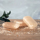 Drei schimmernde, durchscheinende Seifenstücke liegen auf einer Holzoberfläche mit rosa Salzkristallen, neben einem dunklen Olivenzweig.