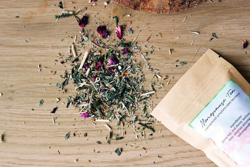Eine Draufsicht zeigt einen Haufen getrockneter Kräuter und rosa Rosenblüten auf einer hellen Holzoberfläche, wobei ein brauner Papierbeutel mit der Aufschrift „Menopause Tee“ teilweise sichtbar ist. Weitere verstreute Kräuter befinden sich um den Haupthaufen herum, was auf ein natürliches und biologisches Produkt hindeutet. Ein Baumstumpf ist oben links zu sehen.