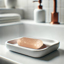 Ein einzelnes, abgerundetes rosa Himalaya-Salzseifenstück liegt in einer minimalistischen weißen Seifenschale am Rand eines Waschbeckens.