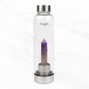  Eine transparente "myga" Wasserflasche mit einem spitz zulaufenden violetten Amethystkristall auf einem silbernen Sockel, stehend auf einer weißen Marmoroberfläche.