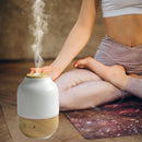 Aroma-Diffusor aus Holz und weißer Keramik, der Dampf abgibt, vor einer in Yoga-Pose sitzenden Person.