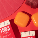 Orangefarbene Seifen und Koji White Kojic Acid-Verpackungen auf einem leuchtend roten Hintergrund, mit strukturierten Elementen und Garn, die Tiefe und Kontrast verleihen.