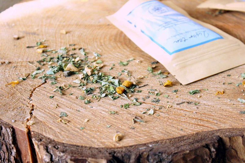 Nahaufnahme einer „Lebertee“-Packung mit blauem Etikett, das die Vorteile für Leber und Gallensystem sowie Inhaltsstoffe wie Löwenzahnblatt und Kurkuma detailliert beschreibt. Teeblätter sind um die Packung auf einer Holzoberfläche verstreut, was ein erdiges und natürliches Gefühl vermittelt.