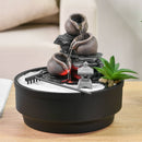 Ein kleiner Zimmerbrunnen mit Steintöpfen und einem Zen-Garten in einer schwarzen Schale.
