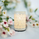 Ein leuchtender weißer elektrischer Wachswärmer mit einem sich wiederholenden, vertikalen Farn- oder Blattausschnittmuster, der auf einer hellblauen Oberfläche steht und von unscharfen weißen und rosa Blüten umgeben ist.