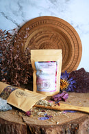  Zwei Teebeutel stehen auf einer Holzoberfläche. Einer ist geöffnet und zeigt lose Teeblätter und getrocknete Blumen, die mit einem Holzgerät aufgesammelt werden. Ein großer geschnitzter Holzteller und getrocknete Blumen sind im Hintergrund zu sehen.