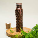 Eine gehämmerte Kupferwasserflasche mit dunklem, sechseckigem Wabenmuster, präsentiert auf einem Holzpodest neben einer brennenden Kerze und grünen Pflanzen.