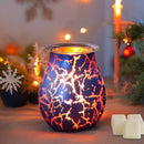 Ein leuchtender elektrischer Wachswärmer aus Glas mit blauer Oberfläche und einem orangefarbenen, feurigen Krakelé-Muster, der auf einem Tisch mit drei Wachswürfeln und festlicher Weihnachtsdekoration steht.