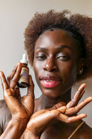 Eine Frau mit Afro-Frisur und strahlender Haut hält eine Flasche Paavani Ayurveda Tridoshic Serum hoch.
