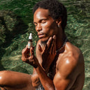 Ein Mann mit dunklen, lockigen Haaren sitzt in klarem grünen Wasser, trägt Gesichtsserum auf seine Wange auf und hält dabei die Pipettenflasche.