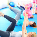 Eine Gruppe von Frauen macht Pilates-Übungen auf blauen Matten, wobei sie pinke Mini-Gymnastikbälle über ihren Köpfen halten.