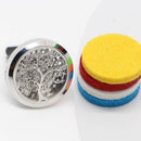 Ein silberfarbener Auto-Aromadiffusor mit einem filigranen ausgeschnittenen Lebensbaum-Design wird neben einem Stapel von vier bunten Ersatz-Filzpads (gelb, rot, weiß und blau) gezeigt.