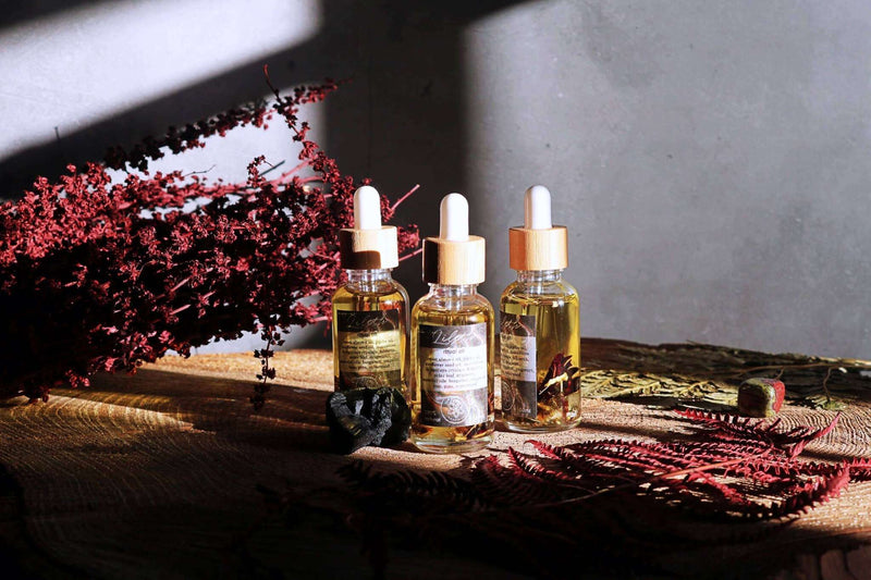 Drei Glasflaschen mit Pipette und Bambusverschluss, gefüllt mit Ritualöl, stehen auf einer Holzoberfläche und werden von Sonnenlicht beleuchtet. Die Flaschen enthalten goldenes Öl und Pflanzenbestandteile. Rote Trockenblumen, ein schwarzer Stein und ein roter Farn sind dekorativ arrangiert, mit einer grauen Wand im Hintergrund.