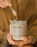 Eine Person mit zart bemalten Fingernägeln schöpft mit einem kleinen Holzlöffel weißes Pulver aus einem Glas "PAAVANI AYURVEDA COCONUT MILK BATH bathing ritual" (240 g), wobei das Pulver beim Schöpfen leicht verstreut wird.