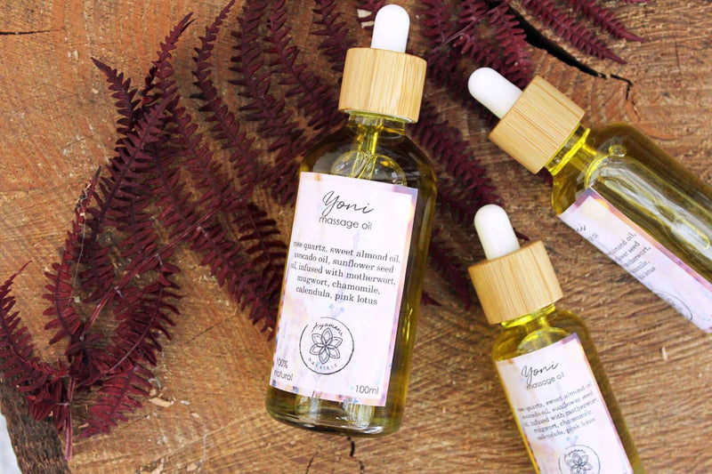 Drei "Yoni Massage Oil"-Flaschen mit hellen Holz-Pipettenverschlüssen sind auf einer Holzoberfläche ausgestellt, die wie ein Baumstamm-Querschnitt aussieht. Die Flaschen variieren in der Größe, und dahinter bildet üppiges grünes und rötliches Laub einen weichen