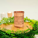 Ein dekorativer Kupfer-Becher oder -Glas mit aufwendigen geprägten Naturmustern, platziert auf einer Holzscheibe und umgeben von grünem Laub, mit zwei unscharfen Kerzen im Hintergrund.