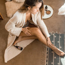 Eine Frau in einem Cardigan sitzt auf einem Teppich und trägt Körperöl auf ihr Bein auf, mit einer Flasche Öl in der Nähe.