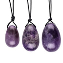 Ein Set aus drei polierten Amethyst-Yoni-Eiern in abgestuften Größen (klein, mittel, groß). Die tiefvioletten, marmorierten Kristalle hängen vertikal an ihren schwarzen Rückholkordeln.