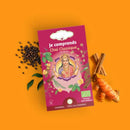 Eine Schachtel "Je comprends Chai Classique" Ayurveda-Tee, umgeben von schwarzem Pfeffer, Kurkuma und Zimtstangen auf orangefarbenem Hintergrund.
