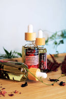 Drei Glasflaschen mit Pipette für "Hot n Spicy Tantric Massage Oil" sind auf einer hellen Holzoberfläche ausgestellt, wobei eine Flasche im Vordergrund auf der Seite liegt. Verstreute getrocknete dunkelrote Blütenblätter und hellere rosa Blütenblätter umgeben die Flaschen, ergänzt durch eine grüne Ranke und einen unscharfen Rosenquarzkristall im Hintergrund.