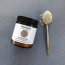 Ein offenes Glas "PAAVANI AYURVEDA ASHWAGANDHA" (3.5 oz / 100g) und ein Teelöffel mit beige-farbenem Pulver liegen auf einer grauen, gesprenkelten Oberfläche, was auf ein natürliches, pflanzliches Ergänzungsmittel hinweist.