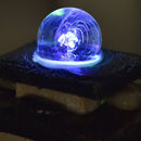 Eine Nahaufnahme einer blau leuchtenden Glaskugel, die auf einem Steinbrunnen rotiert, mit magischen Lichtwirbeln und Glasfasereffekten im Inneren der Kugel.