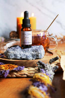 Eine kleine Tropfflasche „Blue Lotus Tincture“ ruht auf einem rauen blauen Kristall. Der Kristall und die Flasche befinden sich auf einem Holztablett mit einem Glas Tee und getrockneten lila Lotusblüten im Vordergrund und brennenden Kerzen im Hintergrund.