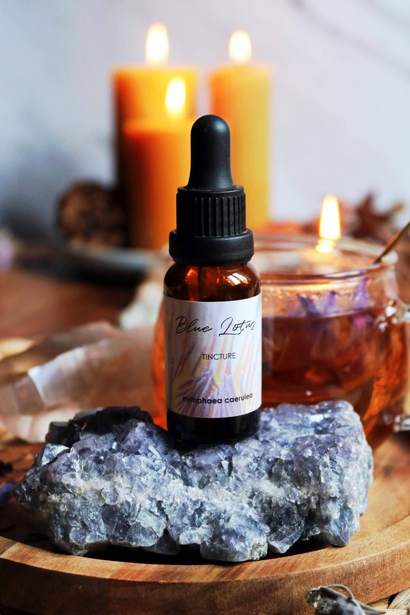 Nahaufnahme einer kleinen Glas-Tropfflasche mit der Aufschrift „Blue Lotus Tincture“, die auf einem Stück rauem lila-blauem Kristall ruht. Im unscharfen Hintergrund sind brennende Kerzen, klarer Quarz und ein Glas mit bernsteinfarbener Flüssigkeit zu sehen.