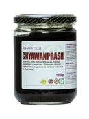 Braunes Chyawanprash in einem Glas mit schwarzem Deckel, auf dem ein Etikett mit dem Namen "CHYAWANPRASH", "500 g" und Symbolen für "100% Natural" und "OMG" abgebildet ist.