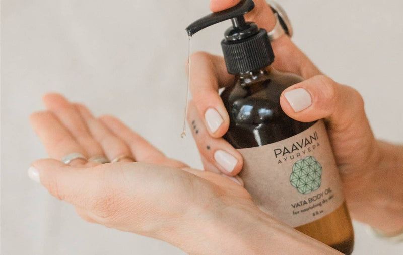 Nahaufnahme von Händen mit hellem Nagellack und Ringen, wobei eine Hand eine PAAVANI Vata Körperölflasche hält und einen Ölstrahl in die gewölbte Handfläche der anderen Hand pumpt.