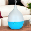 Ein weiß-blauer tropfenförmiger Aroma-Diffusor, der Nebel ausstößt und auf einem Holztisch vor einer weißen Couch in einem Wohnzimmer steht.