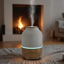 Aroma-Diffusor aus Holz und weißer Keramik mit blauem Licht und Dampf vor einem leuchtenden Kamin.