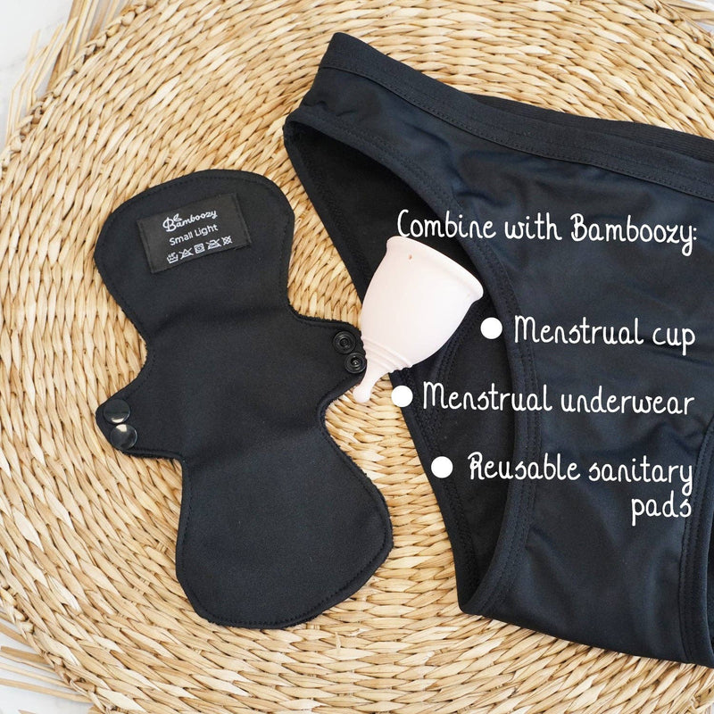 Eine schwarze wiederverwendbare Binde (Small Light), ein hellrosa Menstruationsbecher und schwarze Menstruationsunterwäsche auf einer gewebten Matte, zeigt ein komplettes wiederverwendbares Hygienekonzept von Bamboozy.
