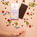 Ein Delikao "Super Woman" Schokoladenriegel und seine Verpackung liegen auf einer rosa Oberfläche, umgeben von verstreuten Mandeln, Himbeeren, blauem Borage und Schokoladentropfen. Eine Hand liegt im Vordergrund.