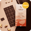 Eine Delikao Skin Glow 70% Zartbitterschokolade mit Brasilnuss und ihre Verpackung, die "Grand Prix Indies Days Cosmétiquemag 2024" auszeichnet, sind zusammen mit Nüssen auf einer hellbraunen Oberfläche abgebildet.