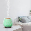 Ein dekorativer, abgerundeter Ultraschall-Aromadiffusor in weißer Lamellenoptik, der limettengrün leuchtet und Dampf abgibt. Er steht auf einem Nachttisch in einer verschwommenen, hellen Schlafzimmereinrichtung.