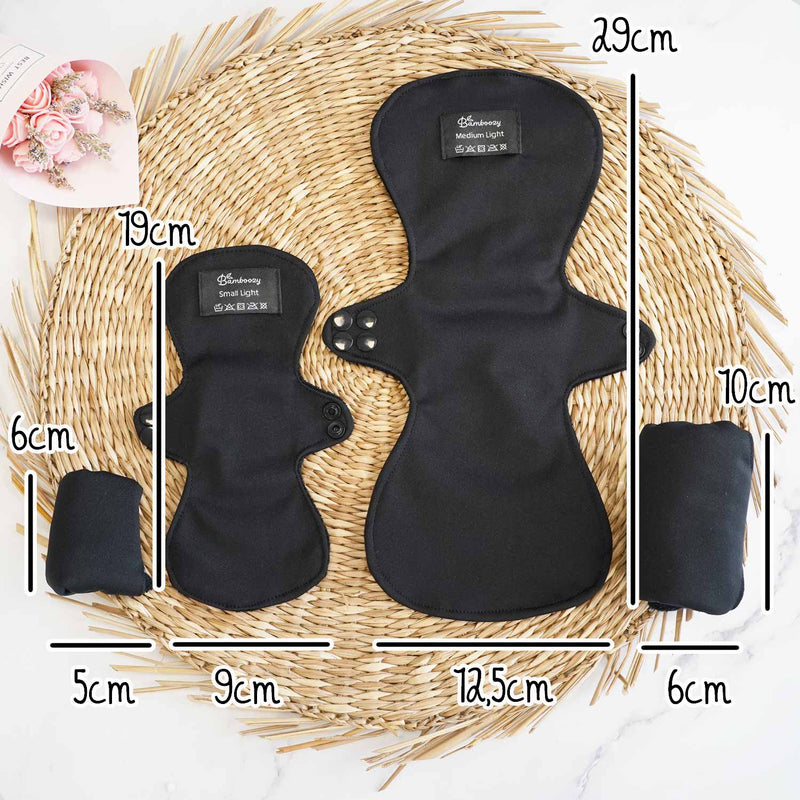 Zwei schwarze wiederverwendbare Binden von Bamboozy (Small Light und Medium Light) auf einer gewebten Matte mit Größenangaben für Binden (19cm, 29cm Länge; 9cm, 12.5cm Breite) und gefaltet (5cm, 6cm Breite; 6cm, 10cm Höhe).