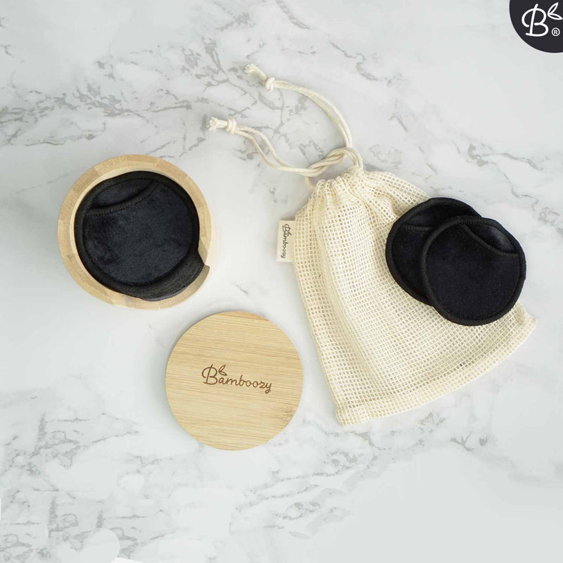 Schwarze wiederverwendbare Make-up-Entfernerpads, ein Bambushalter und ein Wäschenetz von Bamboozy auf einer Marmoroberfläche.