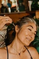  Eine lächelnde Frau mit Sommersprossen und grauen Strähnen trägt mit einer Pipette goldfarbenes Öl auf ihr Ohr auf, wahrscheinlich ein Pflegeprodukt.