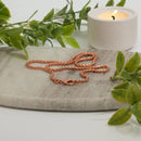 Eine geflochtene Kupferkette mit Verschluss liegt auf einem Marmortablett, neben einer brennenden Kerze und grünen Blättern.