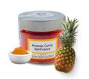 Ein Glas Ananas-Curry-Senf-Sauce mit der Aufschrift „Edition Gold“, daneben eine kleine Schüssel Currypulver und eine frische Ananas, die einen tropischen Geschmack vermitteln.