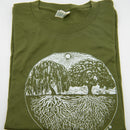 Ein gefaltetes, olivgrünes T-Shirt mit einem großen, kreisförmigen weißen Motiv von zwei kontrastierenden Bäumen—einer Trauerweide, einer voller—deren Wurzeln miteinander verwoben sind.