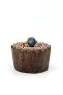 Ein goldener Fingerring mit einem runden, schillernden blaugrauen Labradorit-Edelstein, präsentiert auf einem geschnitzten Holzsockel mit Mandala-Muster.