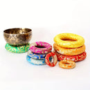 Eine Klangschale ruht auf einem grünen Kissen, umgeben von einem Stapel bunter, gemusterter Kissen.