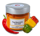 Glas mit orangefarbener Paprika-Chili-Creme und silbernem Deckel mit der Aufschrift „Paprikacreme mit Chili“. Umgeben von bunten roten, gelben und grünen Paprikaschoten. Hell und appetitlich.