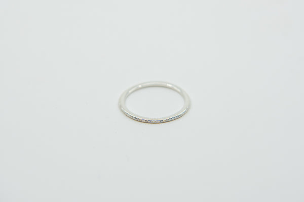 Ein dünner silberner Ring mit einer gemusterten oder texturierten Oberfläche, auf einem weißen Hintergrund.