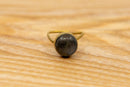 Eine Nahaufnahme aus niedriger Perspektive eines einfachen goldfarbenen Rings mit einem runden, dunkelgrauen Stein in einer Fassung. Der Ring liegt auf einer hellen Holzoberfläche mit sichtbarer Maserung.