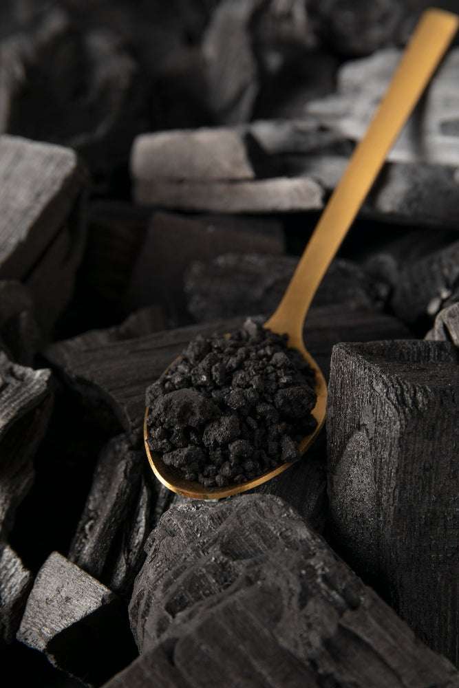 Schwarzes Aktivkohle-Pulver auf einem goldenen Löffel vor Holzkohle-Hintergrund.